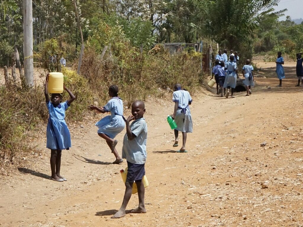 Weltwassertag – Sauberes Wasser statt langer Wege: Ein Kind in Schuluniform trägt einen schweren Wasserkanister auf dem Kopf, während andere Kinder zur Schule gehen. Millionen Kinder in Afrika verbringen täglich Stunden mit dem Wasserholen, anstatt im Unterricht zu lernen.“