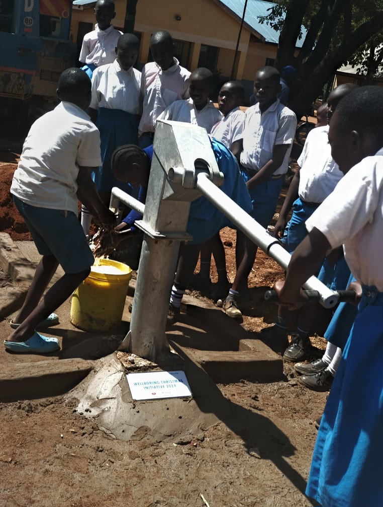 Schüler:innen der Kodero Bara Primary School in Afrika stehen um ihren neuen Brunnen mit Namensplakette – ermöglicht durch die WellBoring Christmas Initiative 2024. Zugang zu sauberem Trinkwasser verbessert Gesundheit und Bildung.