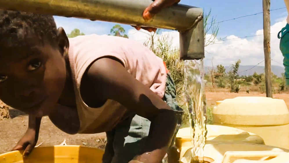 Ein Kind an einem WellBoring Brunnen in Afrika macht Spass mit der Kamera, während frisches Wasser aus der Pumpe strömt. Dank Projekten wie WellBoring erhalten immer mehr Gemeinden sicheren Zugang zu sauberem Trinkwasser. Brunnenbau in Afrika verändert Leben – Weltwassertag erinnert daran, wie wichtig Wasser für die Zukunft ist.