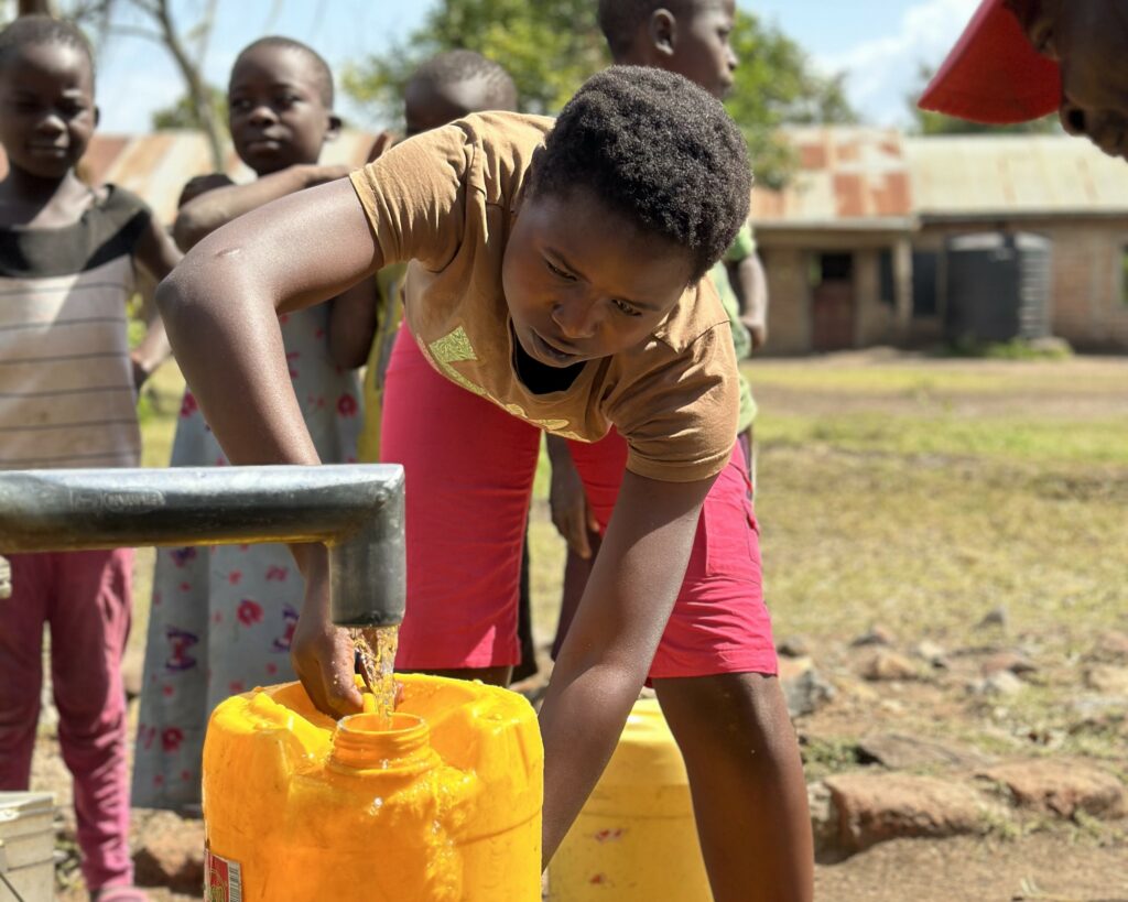 Ein junges Mädchen in Afrika füllt einen Kanister mit sauberem Wasser aus einem WellBoring-Brunnen auf dem Schulgelände. Im Hintergrund warten weitere Kinder darauf, frisches Trinkwasser zu holen.