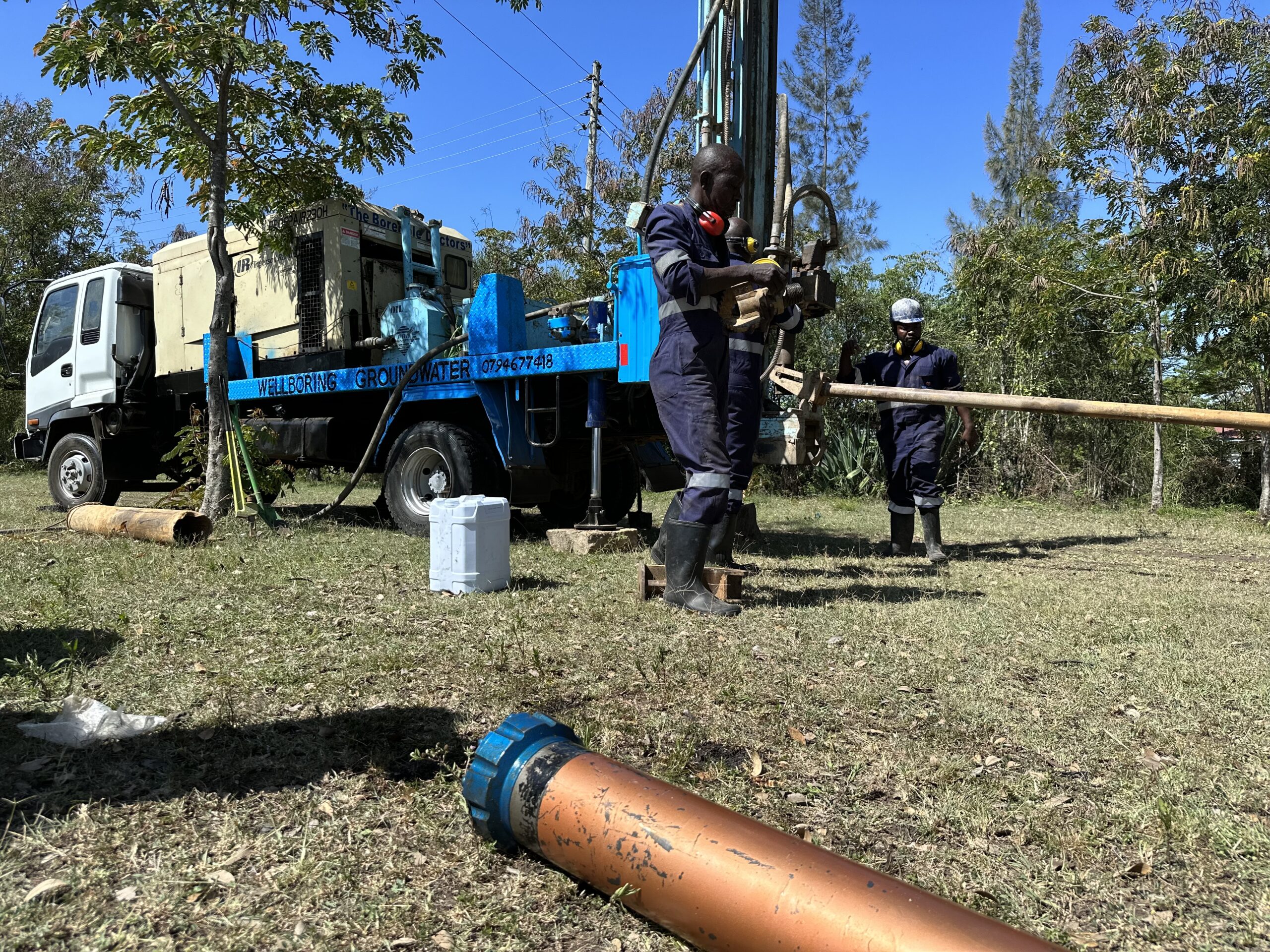 Expert drilling team members from WellBoring methodically add an extension to a drill bit, part of the sustainable water well construction process in a grassy field.