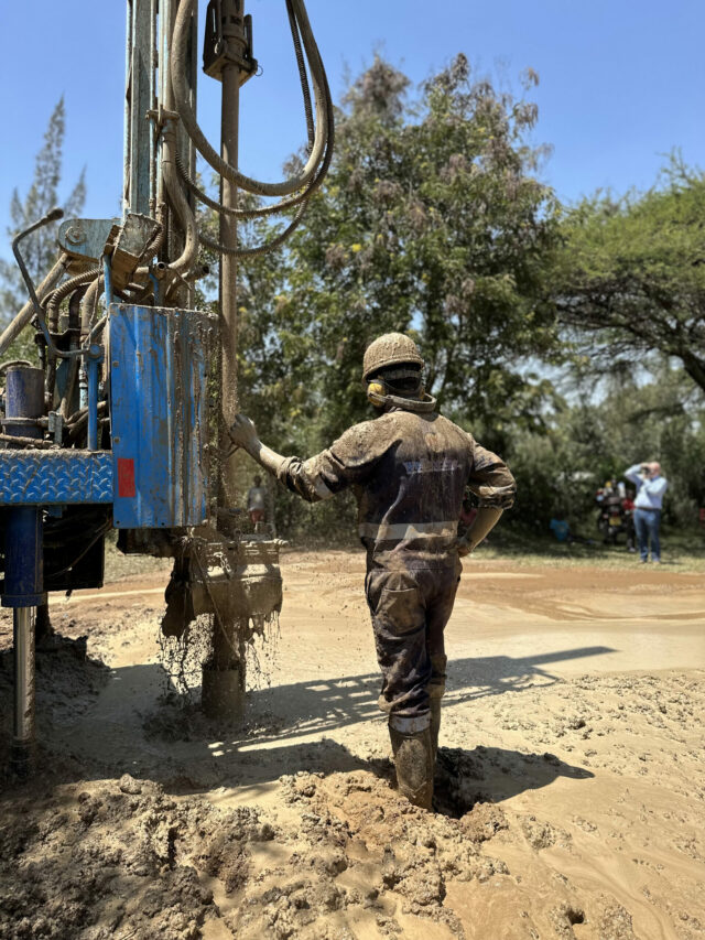 A WellBoring Groundwater worker, covered in mud, stands beside a drilling rig, a sign of successful water finding during a well-drilling operation.