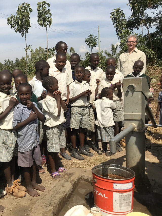 School children in uniforms gathered around a new hand pump well in their schoolyard, eagerly awaiting fresh water.
