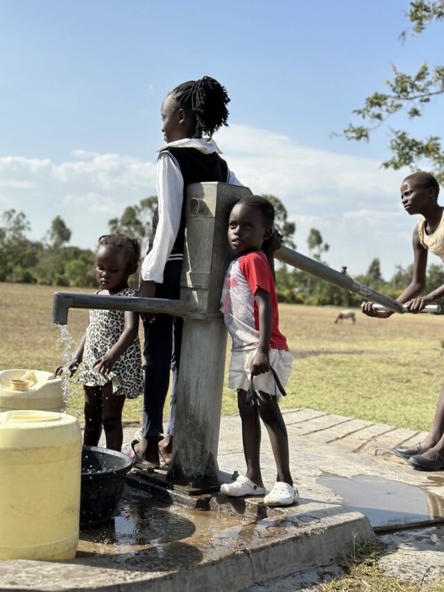 Children gather around a wellboring water pump at a school, a symbol of positive change and assistance for schools in critical need.