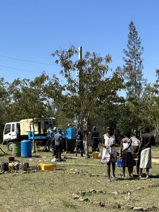 WellBoring's drilling team works at a Kenyan primary school, observed by three community members, symbolizing progress towards secure water access.