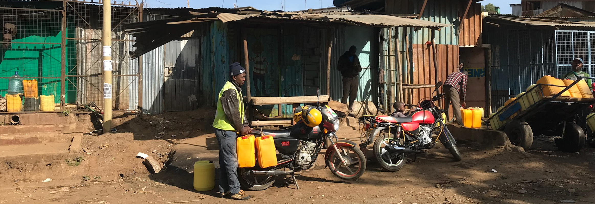 A man in a high visibility jacket carefully balances yellow jerry cans filled with water on his motorcycle in a bustling community setting, showcasing daily water collection routines.