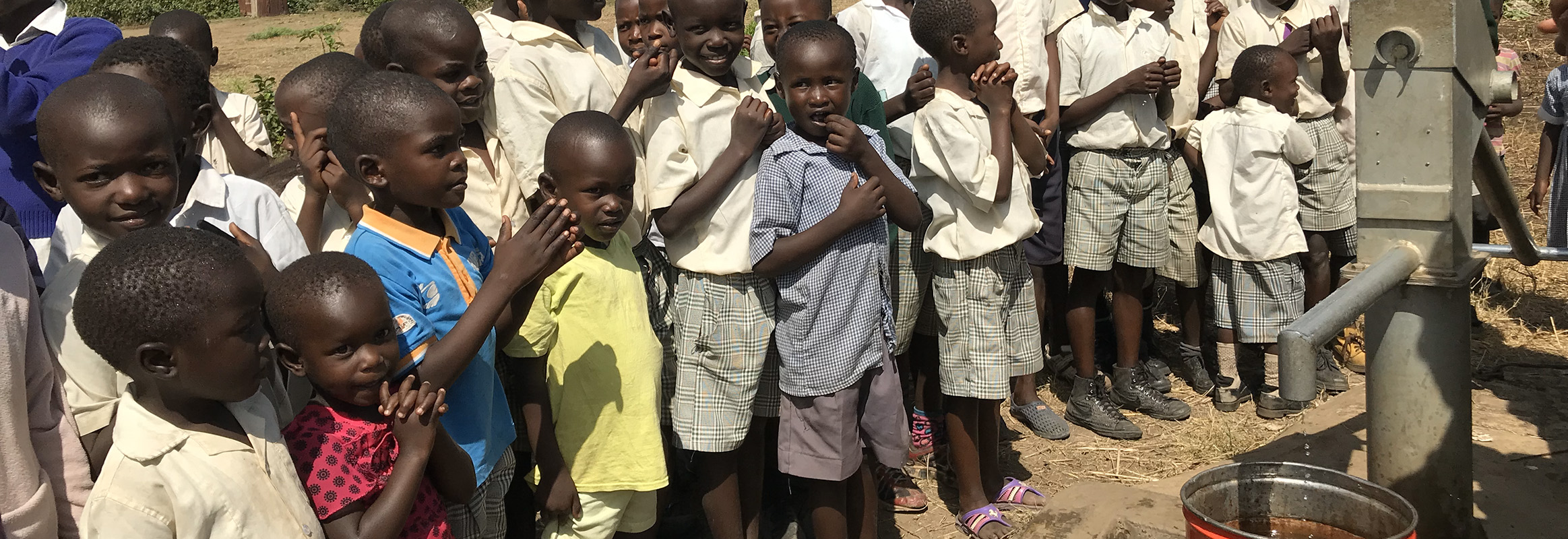 School children in uniforms gathered around a new hand pump well in their schoolyard, eagerly awaiting fresh water.