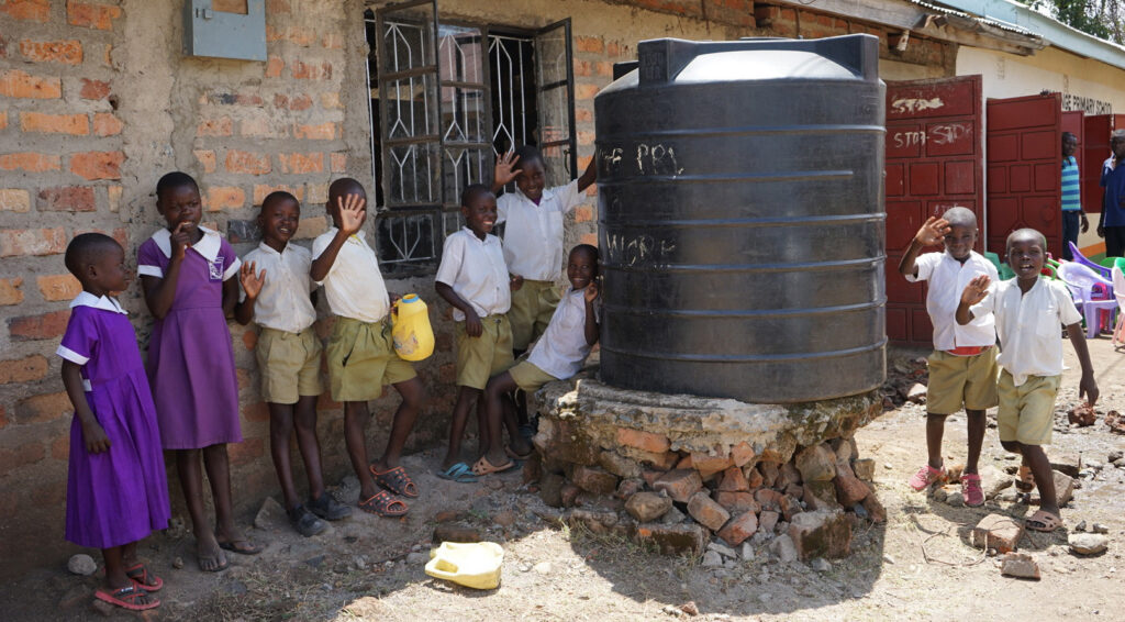 Happy children wave and smile beside a large water storage tank at their primary school, signifying WellBoring's impact on water accessibility.