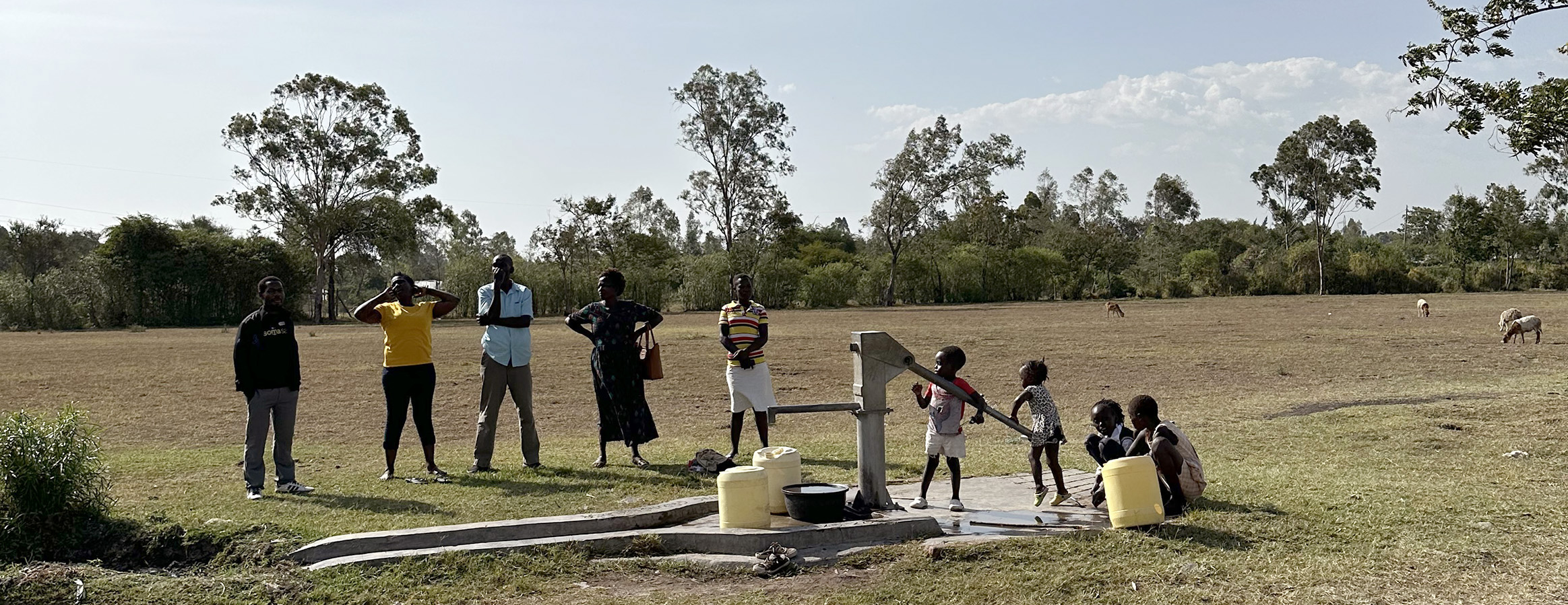 Teachers and a community board member with children gather around a WellBoring water pump in a rural African landscape.